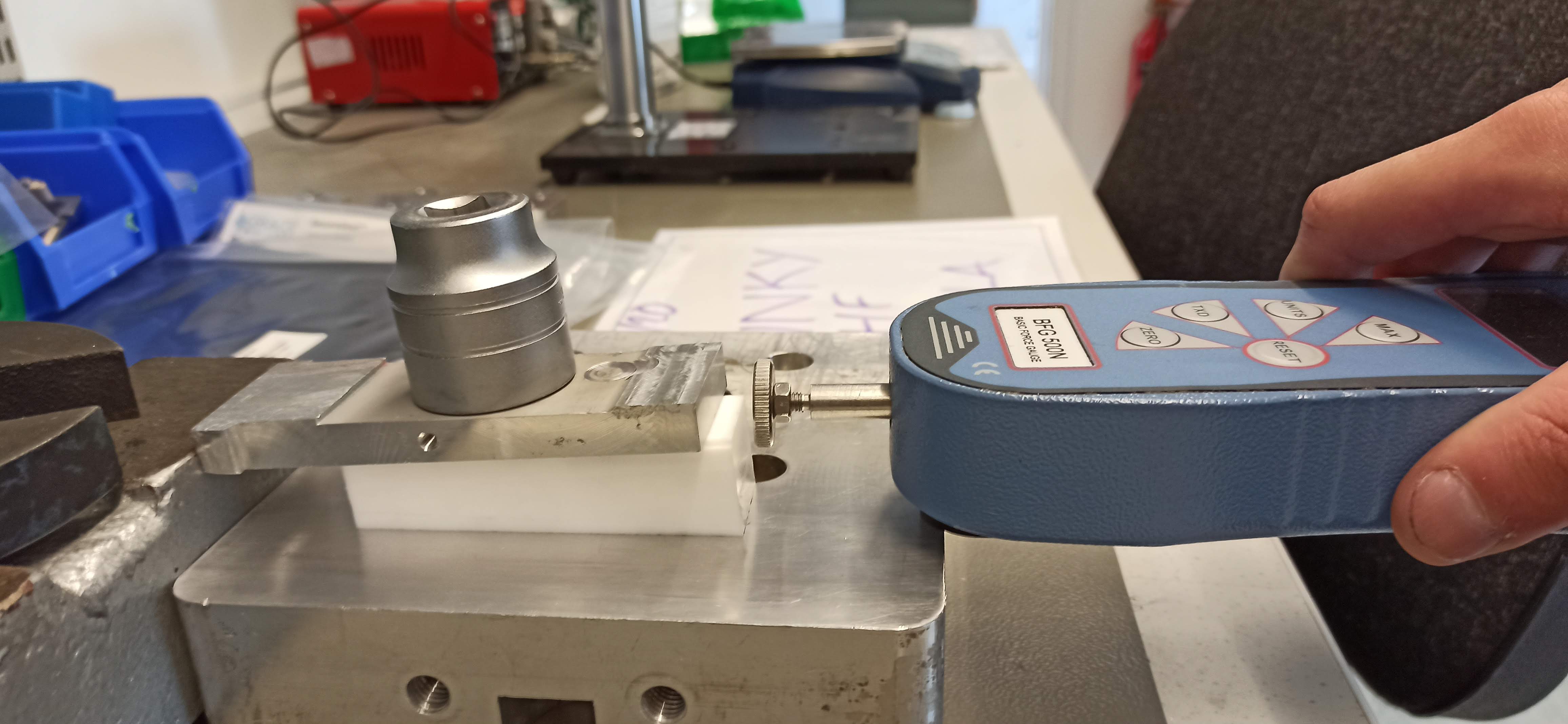 Hand holding a blue force gauge pressing on machined aluminium and white plastic parts clamped in a test fixture, workshop bench behind