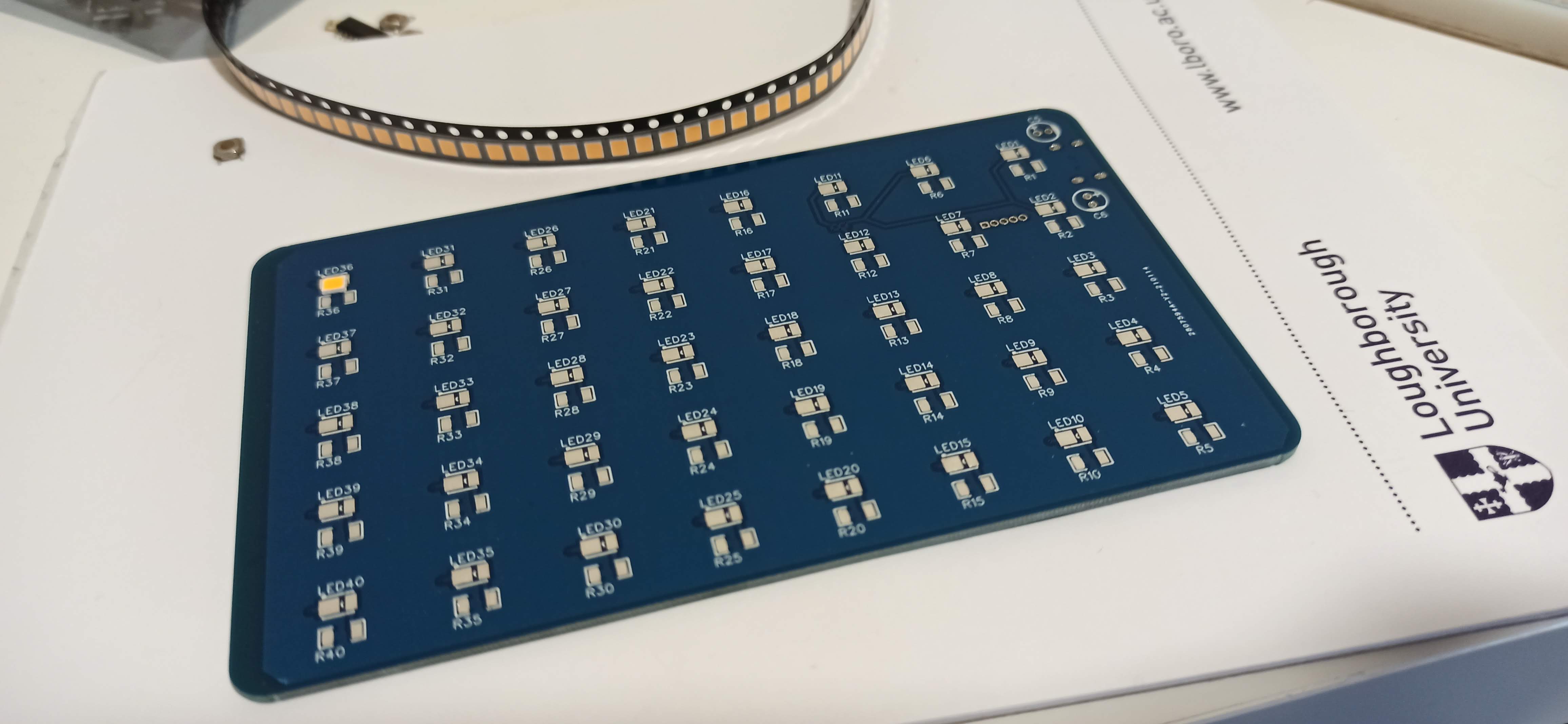 Blue printed circuit board with forty labelled LEDs and resistors, one LED soldered, lying on Loughborough University letterhead beside an LED reel